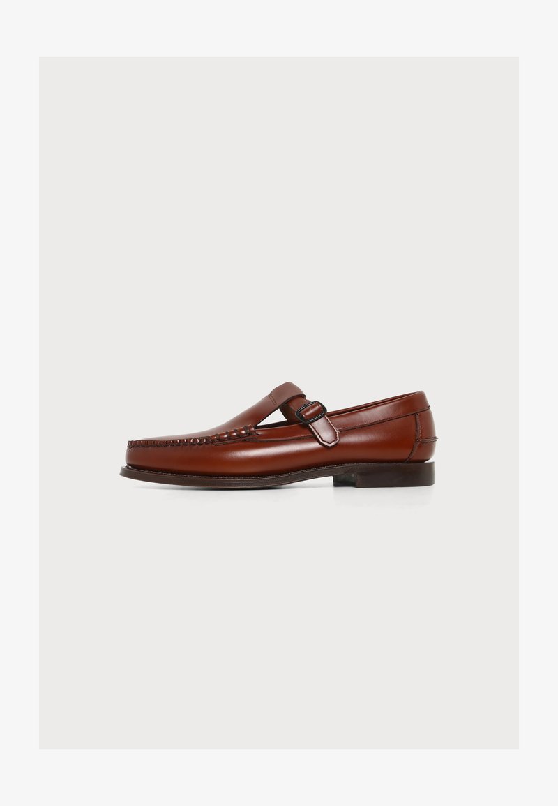 Mocassin marron en cuir avec une bride à boucle et des coutures détaillées à l'avant, présenté sur un fond clair uni.