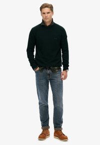 Dunkelgrüner Strickpullover, über einem karierten Hemd getragen, kombiniert mit blauen Jeans und braunen Schnürstiefeln. Glatte Textur, lässige Passform und hochgekrempelte Ärmel.