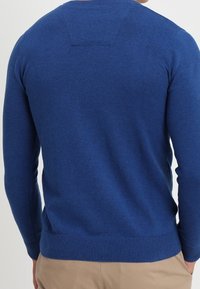 Blå stickad tröja med långa ärmar, ribbad ärmslut och nederkant, med en rektangulär detalj på nedre delen av ryggen. Mjuk textur.