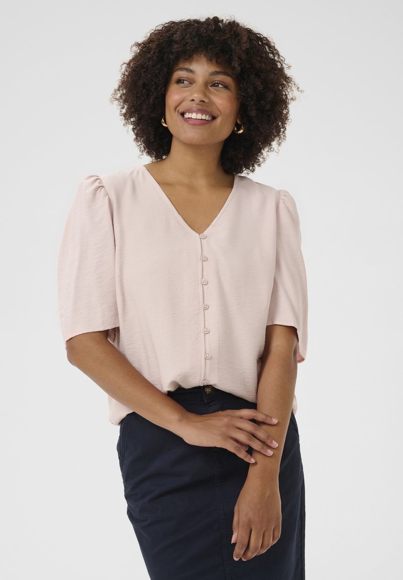 Blusa rosa chiaro con scollo a V, maniche corte a sbuffo e chiusura centrale con bottoni. Tessuto morbido, abbinata a una gonna blu navy.