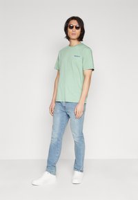 Mintgrønn t-skjorte med korte ermer og logo, kombinert med lyseblå jeans og hvite joggesko, aksessorisert med runde solbriller.