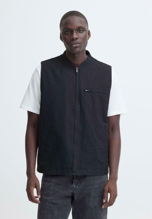 Jonge man die een wit, kort mouwig overhemd draagt onder een zwart gilet met rits en donkere spijkerbroek, staand tegen een effen lichte achtergrond.