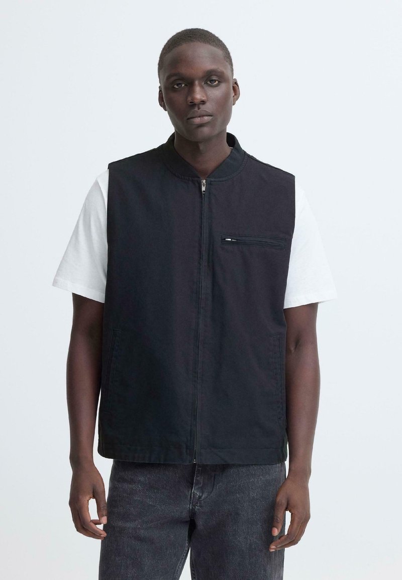 Jeune homme portant une chemise blanche à manches courtes sous un gilet noir zippé et un jean foncé, debout devant un fond clair uni.