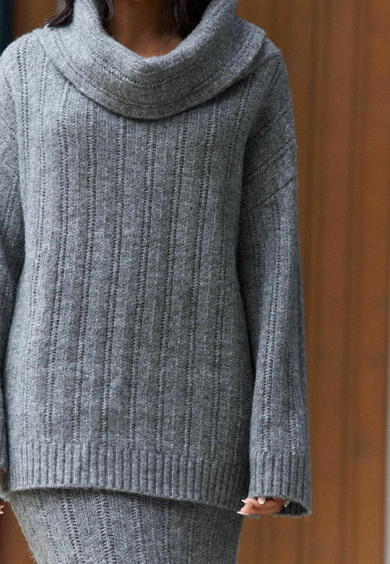 Personne portant un pull en maille côtelée gris avec un large col bénitier et une jupe grise assortie, debout devant un fond en bois.