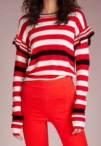 Pull rayé rouge et blanc avec accents noirs, doté de manches à volants et de poignets longs. Associé à un pantalon rouge taille haute.