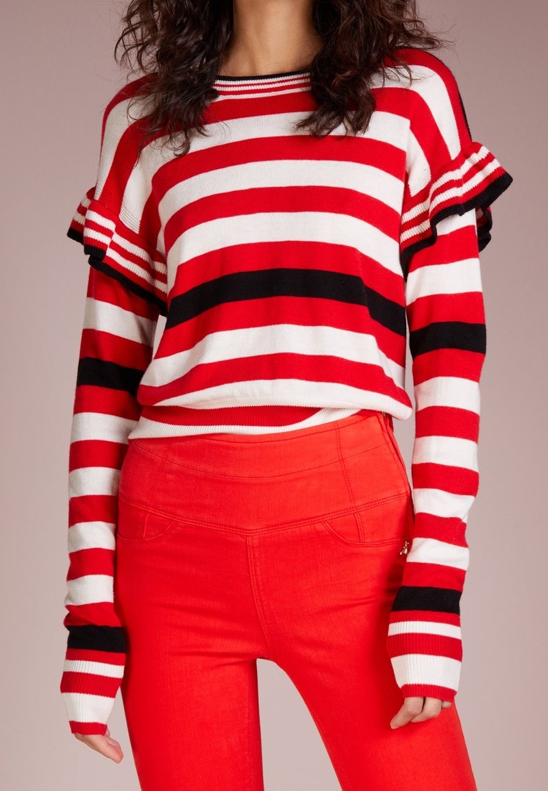 Pull rayé rouge et blanc avec accents noirs, doté de manches à volants et de poignets longs. Associé à un pantalon rouge taille haute.