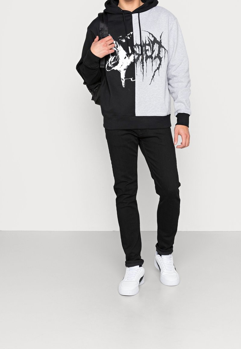 Personne portant un sweat à capuche noir et gris avec un motif graphique blanc représentant un animal rugissant, un pantalon noir, des baskets blanches et un sac à dos noir.