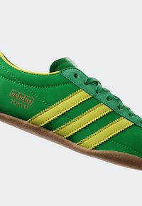 Πράσινα και κίτρινα αθλητικά παπούτσια Adidas Tokyo με τρεις ρίγες, υφασμάτινη επάνω επιφάνεια, καουτσούκ σόλα και υφή στις πλευρές.