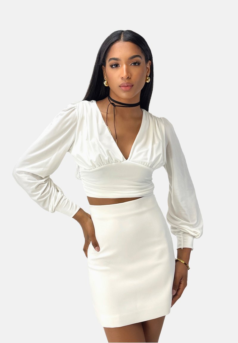 Blusa blanca, corta, con mangas largas abullonadas y un escote en V pronunciado, combinada con una mini falda blanca. Tejido suave y liso con una silueta ajustada.