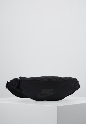 Sort Nike bæltetaske med lynlåslukning og justerbar rem vist på en hvid piedestal mod en ensfarvet grå baggrund.