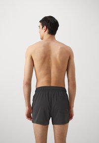 Maillots de bain gris foncé avec une taille élastique et une poche arrière ; le tissu semble lisse et léger, conçu pour les activités aquatiques.