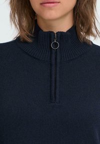 Námornícky modrý pletený pulóver s vysokým golierom a polovičným zipsom, s hladkou textúrou a jemnými rebrovanými akcentmi.