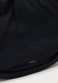 Musta laukku, joka on valmistettu sileästä kankaasta ja jossa on pehmeä pinta. Laukussa on pyöreä muoto ja alhaalla kullattu "SANDQVIST"-logo.