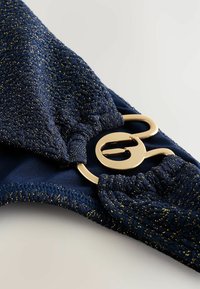 Navy blauwe bikini top gemaakt van textuurrijke glanzende stof, met een gouden ronde sluiting en geplooide details aan de zijkanten.