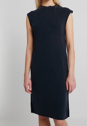 Robe noire sans manches, longueur au genou, à col haut, portée par une personne debout devant un fond uni.