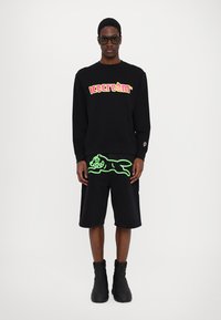 Svart hettegenser med lys rosa "iskrem" logo, parret med svarte shorts med grønn kattedesign. Stylet med svarte ankelsko.