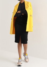 Cappotto giallo doppiopetto su una maglia a righe nera, abbinato a una gonna a tubino nera con spacco frontale e sneakers grigie con dettagli gialli.