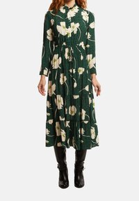 Grön blommig klänning med nedknäppt krage, långa ärmar och volang i nederkant. Har stora vita blommor och en texturerad tyg.