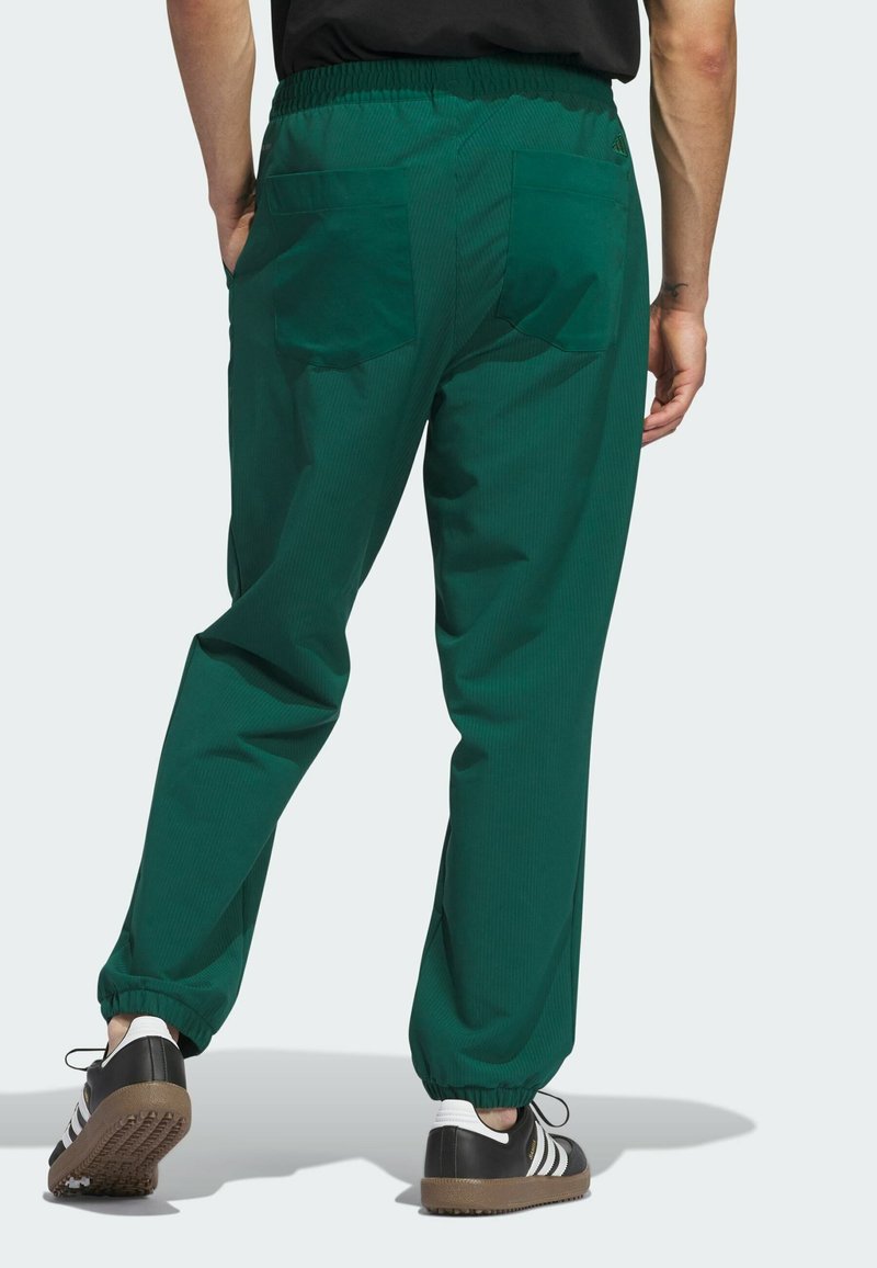 Mann in dunkelgrünen Hosen mit elastischen Bündchen und schwarzen Sneakers mit weißen Streifen, stehend vor einem hellen Hintergrund, die Hände in den Taschen.