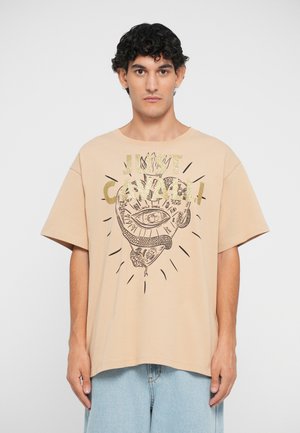 Camiseta de algodón beige con un diseño gráfico en negro que presenta un ojo y una serpiente, acentuado por texto metálico dorado. Corte holgado y mangas cortas.