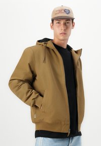 Bruine ritsjack met een capuchon, voorzien van zijzakken en ribgebreide manchetten. Gedirigeerd over een zwart shirt, met lichtblauwe denim eronder.