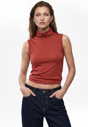 Jonge vrouw met lang haar die een roestkleurige mouwloze coltrui en donkere jeans draagt, handen in de zakken, staand tegen een witte achtergrond.
