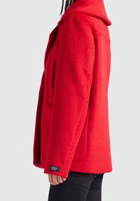 Vue de profil d'une personne portant un manteau en laine rouge vif avec un large col et des poches plaquées, associé à un pantalon noir.