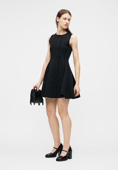 Γυναίκα με μαύρο αμάνικο φόρεμα σε στυλ καμπάνα, που κρατά μια μικρή μαύρη τσάντα με φούντες και φορά μαύρες γόβες με λουράκια στον αστράγαλο.