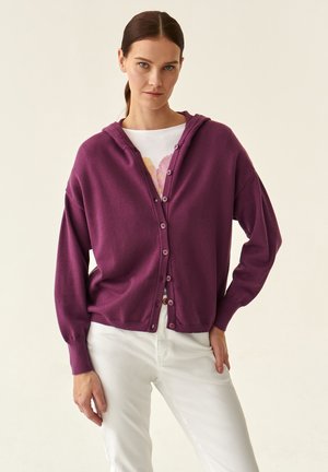 Cardigan viola con cappuccio, chiusura con bottoni, polsini a coste e taglio morbido, indossato sopra una camicia bianca e abbinato a pantaloni bianchi.