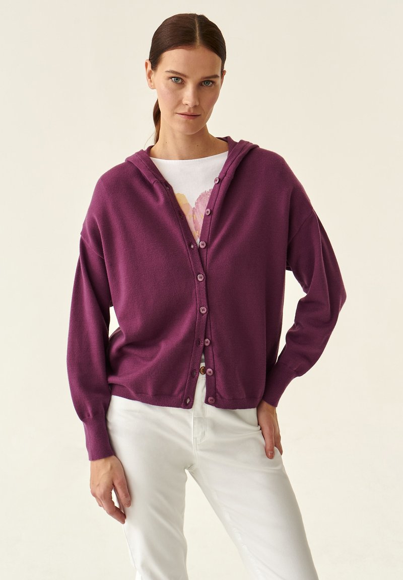 Cardigan viola con cappuccio, chiusura con bottoni, polsini a coste e taglio morbido, indossato sopra una camicia bianca e abbinato a pantaloni bianchi.