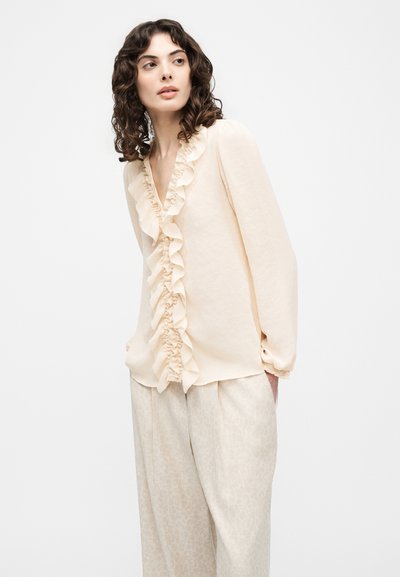 Femme aux cheveux bouclés portant un chemisier écru à volants et un pantalon ample à motif clair se tenant devant un fond uni.