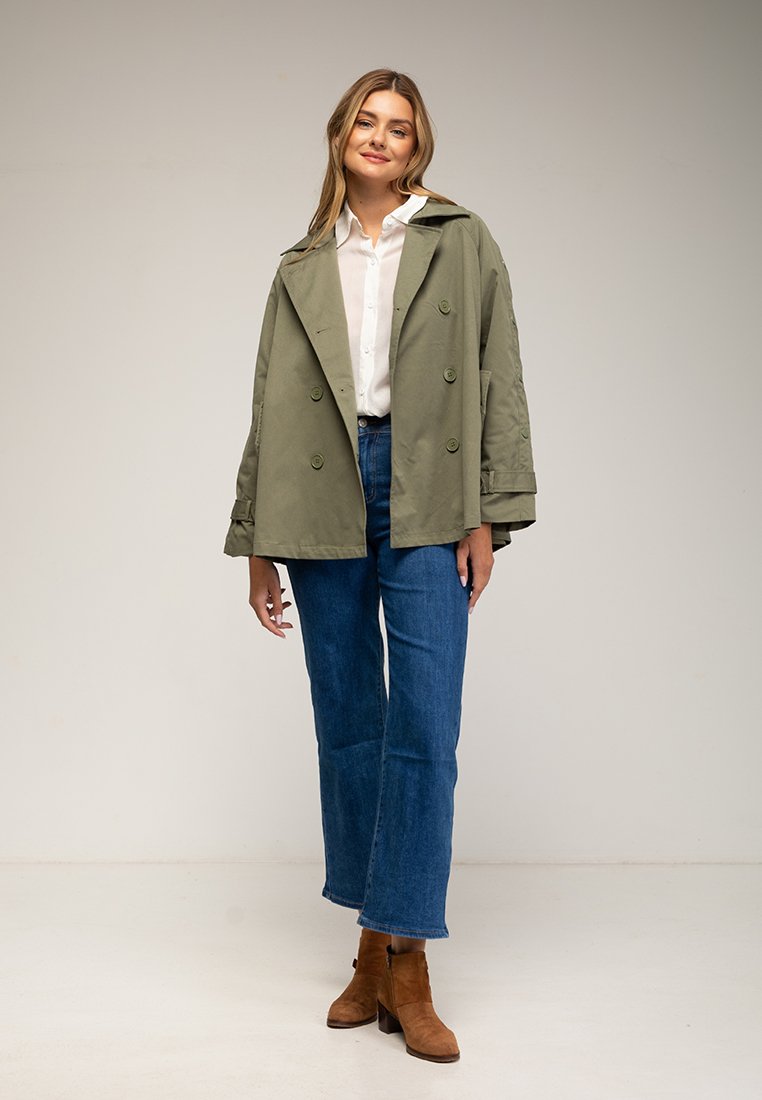 Olivenfarbener, übergroßer Trenchcoat, doppelt knöpfbar mit großen Knöpfen, kombiniert mit blauen weit geschnittenen Jeans und braunen Stiefeletten.