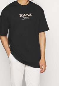 Černé bavlněné tričko s krátkými rukávy a kulatým výstřihem. Na přední straně je potisk loga v světle růžové barvě s textem pod ním. Jednoduchý, nezdobený design.