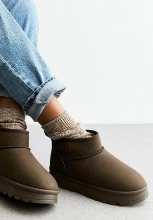Bruine enkellaarsjes van zacht materiaal, met een ronde neus en een gestructureerde zool, gecombineerd met beige geribde sokken en opgerolde lichtblauwe jeans.