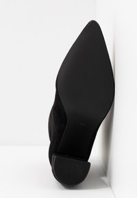 Chaussure noire pour femme à bout pointu, semelle lisse et petit talon, positionnée à la verticale contre des murs blancs, taille 37 visible sur la semelle.