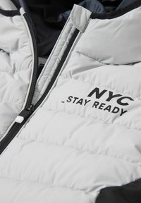 Jasnoszara kurtka puchowa z czarnym zamkiem, czarną podszewką i napisem "NYC STAY READY" wydrukowanym na czole w kolorze czarnym.