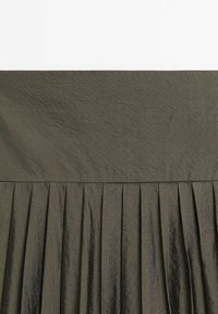 Tissu vert olive foncé avec une large ceinture et de fines plis verticaux sous la ceinture, montrant des rides et une texture.
