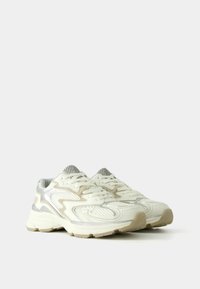 White sneakers with mesh and smooth synthetic overlays, featuring wavy design lines, textured details, and light grey accents on the sole.