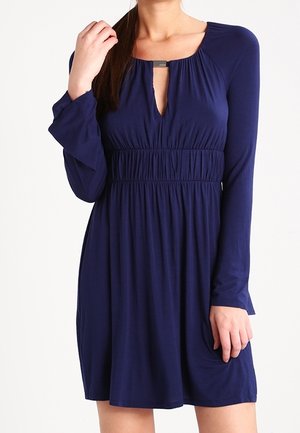 Woman wearing a navy blue long-sleeve dress with a gathered waist and keyhole neckline with metal detail.