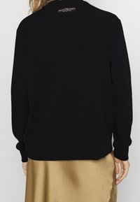 Sudadera negra con cuello redondo y mangas largas, que presenta un dobladillo y puños acanalados, y una pequeña etiqueta con logo en la parte posterior del cuello.