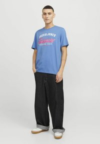 Lyseblå T-shirt med korte ærmer og grafisk tryk i rød og hvid, parret med løse mørkeblå denimjeans og hvide sneakers med brune såler.