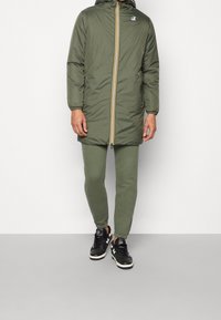 Parka verde lunga con zip e accenti gialli, abbinata a leggings verdi coordinati e sneaker nere, caratterizzata da tessuto strutturato.