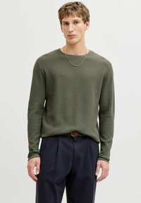 Young man wearing an olive green textured sweater, dark trousers with a brown belt, and a silver chain necklace, standing against a plain background.