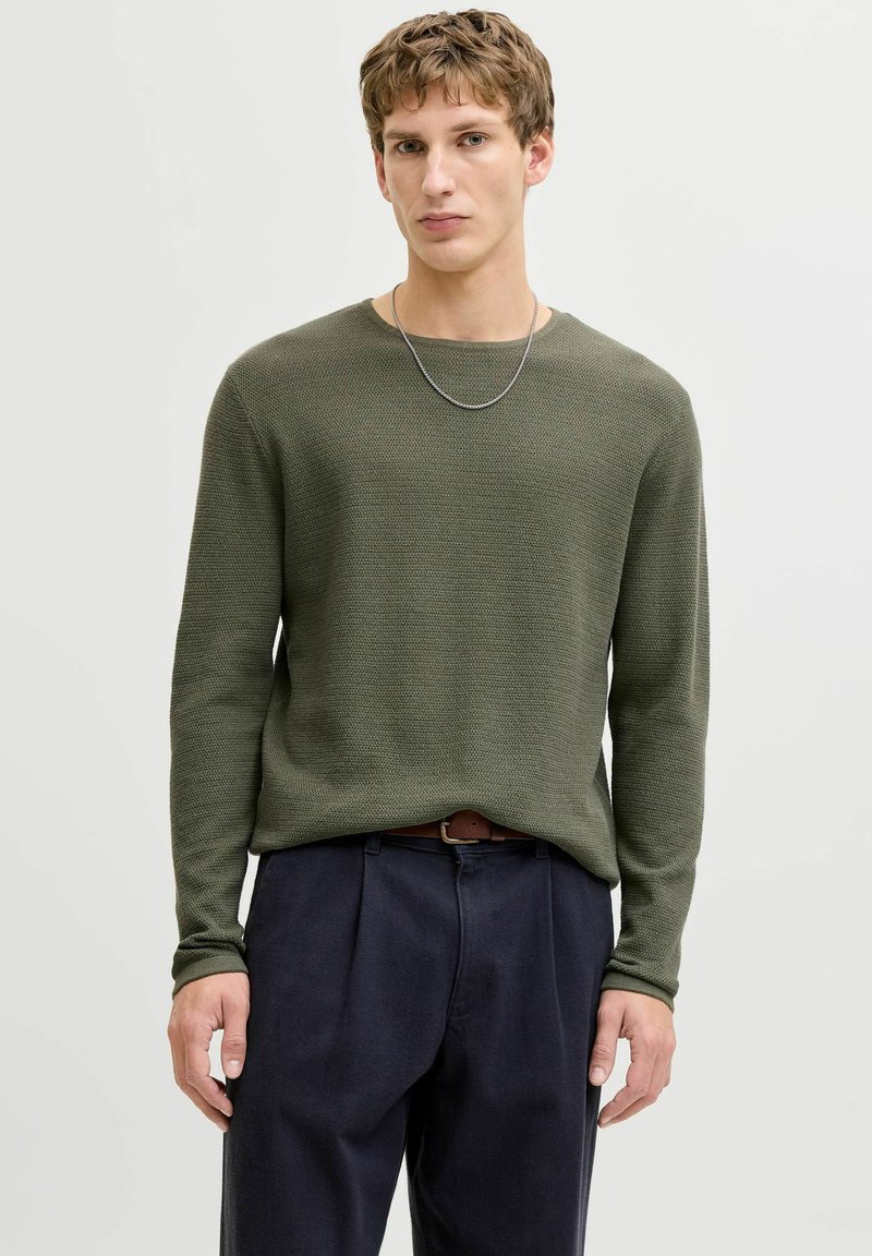 Young man wearing an olive green textured sweater, dark trousers with a brown belt, and a silver chain necklace, standing against a plain background.