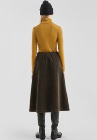 Vrouw in een zwarte muts, mosterdkleurige lange mouw coltrui, donkerbruine corduroy midi-jurk en zwarte kniehoge laarzen, staand met haar rug naar ons toe.