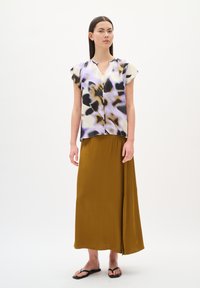 Blouse multicolore avec imprimé floral ; manches courtes et décolleté en V. Une longue jupe jaune moutarde en tissu lisse avec une fente sur le côté. Sandales plates.