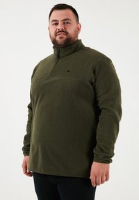 Mørkegrøn fleece-pullover med halv-zip krave, som har en horisontal søm og et lille sort logo på brystet. Blødt materiale.