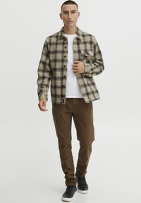 Chemise en flanelle à carreaux marron et noir, avec un t-shirt blanc à col rond en dessous, associée à un pantalon marron et des baskets noires.