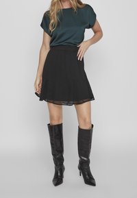 Jupe noire à design plissé, ourlet transparent superposé, associée à un chemisier vert foncé à manches courtes et des bottes en cuir noires montant jusqu'aux genoux.