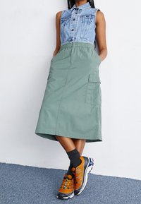Bezrekawnik z denimu z zapięciem na guziki, zestawiony z oliwkowozieloną spódnicą do kolan z kieszeniami i elastycznym pasem, uzupełniony pomarańczowymi tenisówkami.
